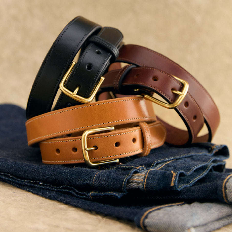 Three leather belts in black, tan, and brown, each with a gold-tone buckle, arranged in a stack on top of folded blue jeans against a neutral beige background.