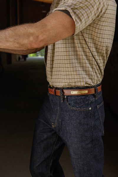 A man wearing a brown leather belt with a gold-tone metal buckle and a beige-and-white checkered short-sleeve shirt, paired with dark blue jeans.