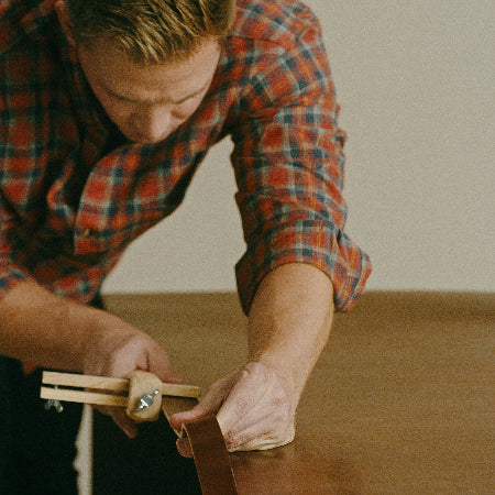 A man in a red and blue plaid shirt uses a wooden measuring tool to mark a straight edge on a wooden table against a plain beige wall.